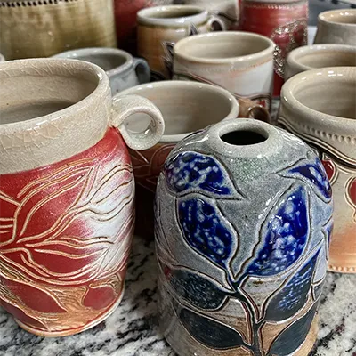 a large selection of soda-ash fired mugs and vases, with brilliant blue and red flower designs carved into their surfaces